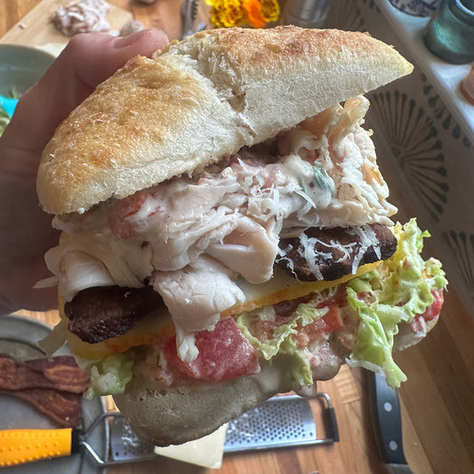 Hand holding a large sandwich with turkey, bacon, cheese, creamy tomato cabbage slaw. with chef knife and cutting board in background