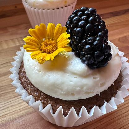 Cupcake with white frosting, yellow flower, and blackberries on a wooden surface