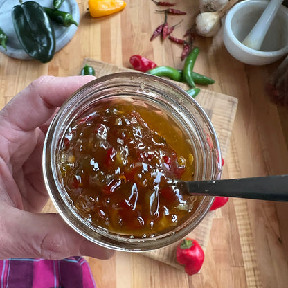 Hand holding a jar of jam with a spoon, surrounded by peppers and a mortar and pestle on a wooden surface.