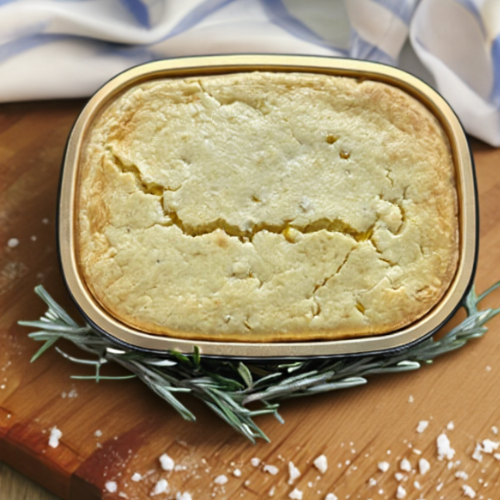 spoonbread in a takeout container on a wooden board with rosemary and salt